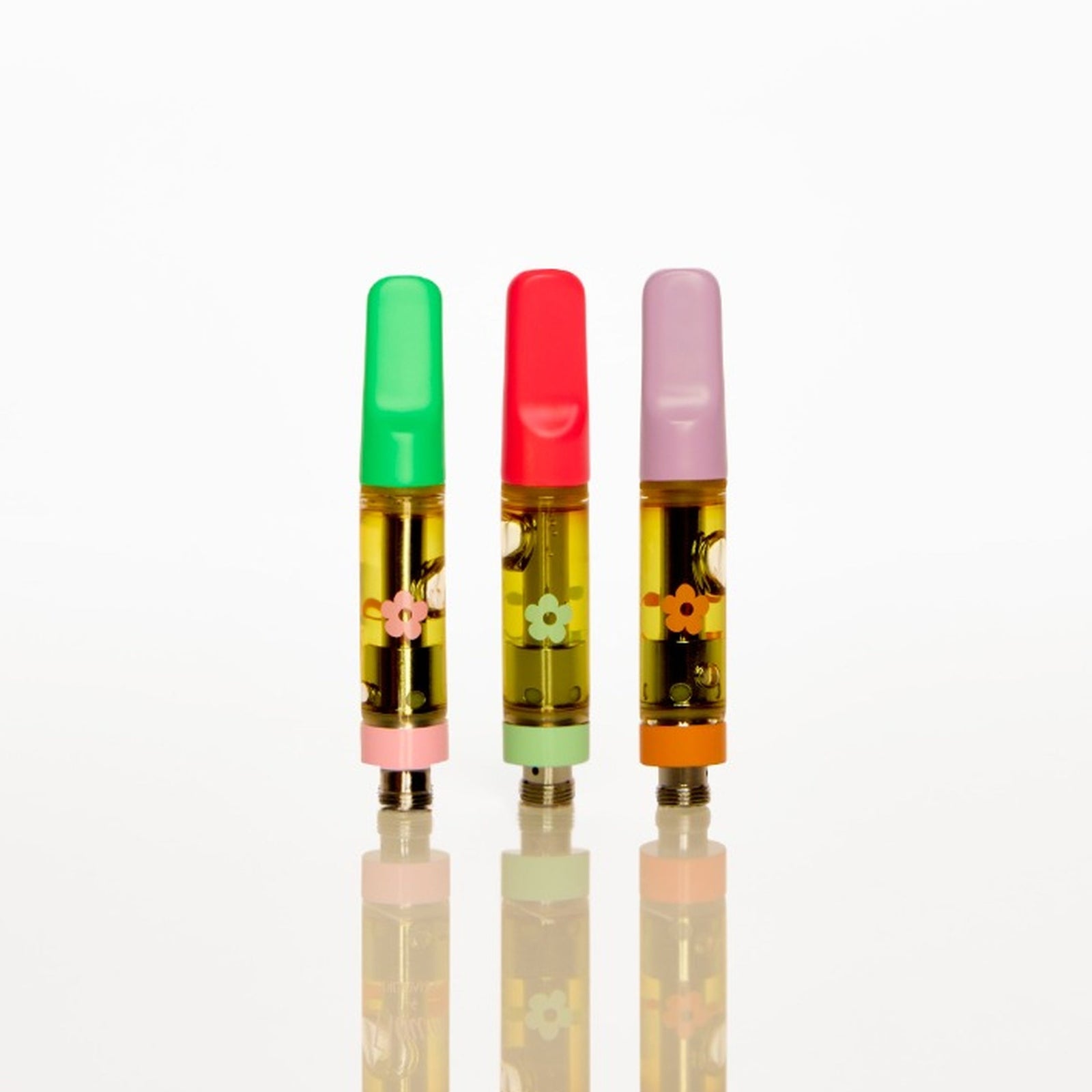 Three small cylindrical vape cartridges standing upright on a reflective white surface against a white background. The left cartridge has a green cap, the middle has a red cap, and the right has a pink cap, all filled with yellow oil and decorated with small floral illustrations.