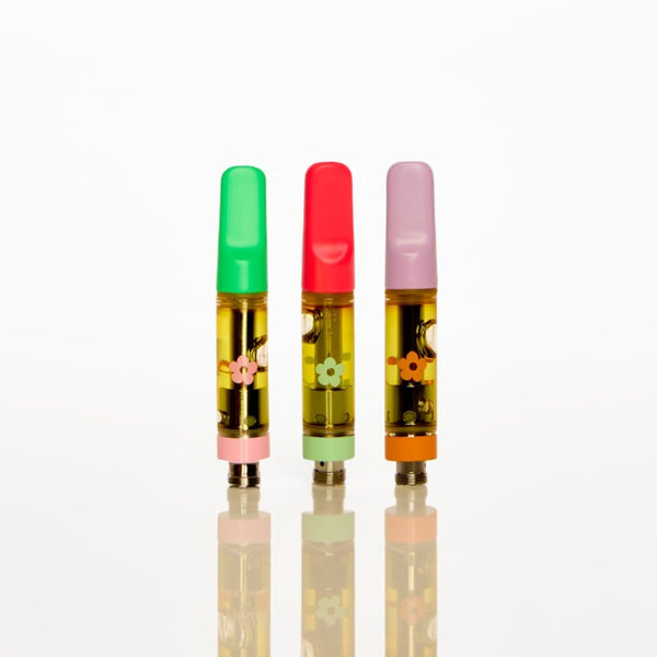 Three small cylindrical vape cartridges standing upright on a reflective white surface against a white background. The left cartridge has a green cap, the middle has a red cap, and the right has a pink cap, all filled with yellow oil and decorated with small floral illustrations.